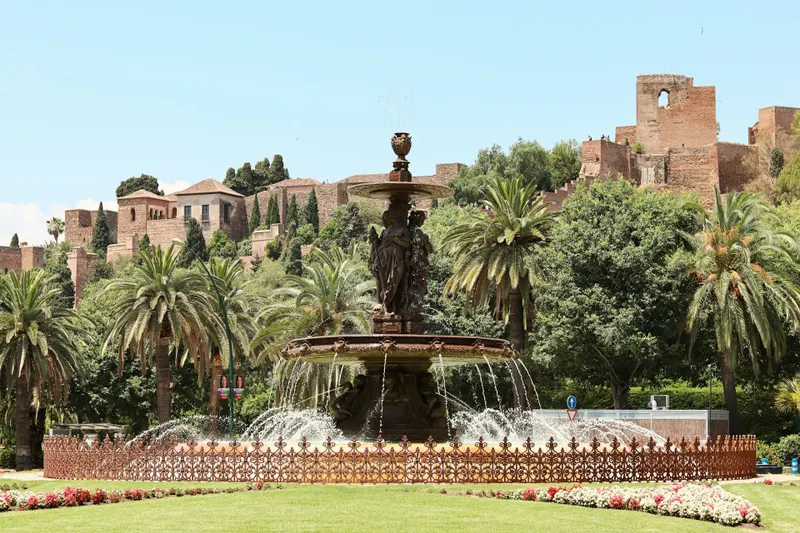 Η σιντριβανάτη Plaza del General Torrijos στη Μάλαγα, με φόντο το επιβλητικό Αλκαζάμπα, περιτριγυρισμένη από φοίνικες, πολύχρωμα λουλούδια και ιστορική αύρα που θυμίζει την αραβική κληρονομιά της πόλης.