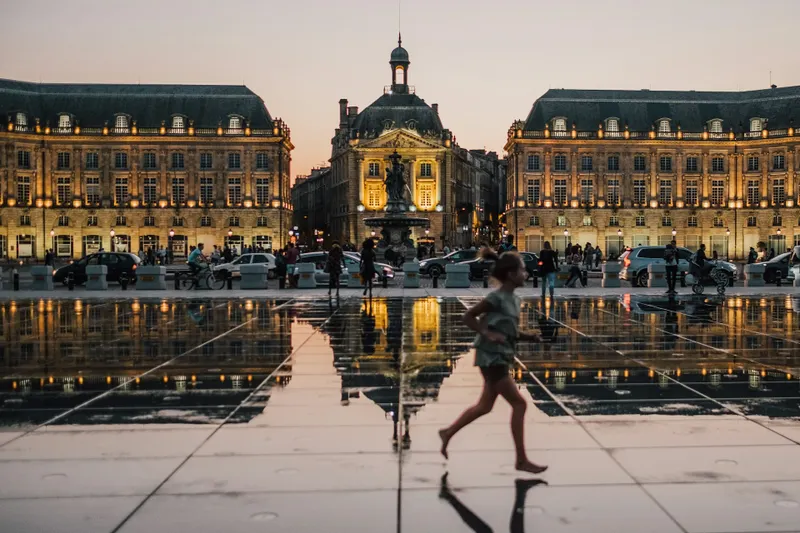 Η Πλατεία Place de la Bourse στο Μπορντό τη στιγμή του δειλινού, με φωτισμένα κλασικά κτίρια να αντανακλώνται στην επιφάνεια του Miroir d'eau, ενώ κόσμος περπατά και ένα παιδί τρέχει στο προσκήνιο.